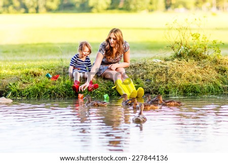 Adorable little girl and her mom playing with paper boats and feeding ducks in a river. Creative leisure with kids.