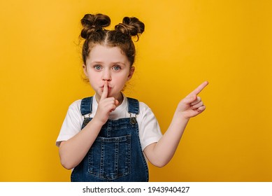 Adorable little girl holding finger on lips symbol of hush gesture of asking to be quiet, isolated over yellow studio background with copy free space for advertisement. Silence or secret concept