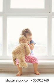 Adorable Little Girl Hugging A Teddy Bear. Cute Baby At Home In White Room Is Sitting Near Window. 