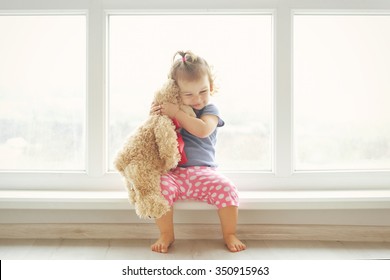 Adorable Little Girl Hugging A Teddy Bear. Cute Baby At Home In White Room Is Sitting Near Window. 