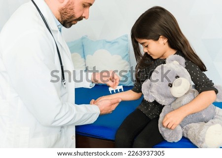 Adorable little girl hugging a teddy bear getting a skin prick test for her allergies with a pediatrician doctor
