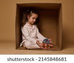 Adorable little girl in pajamas sitting inside a cardboard box and plying with a small colorful house buils from magnetic construction blocks.