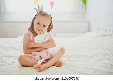 Adorable little girl in pink dress and golden crown dressed as princess playing with unicorn. Kid having fun with soft toy. Child playing with stuffed toy