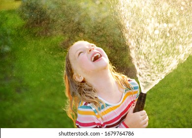 Adorable Little Girl Playing With A Garden Hose On Warm Summer Day. Child Having Fun With Water On Hot Day. Outdoor Summer Activities For Kids.