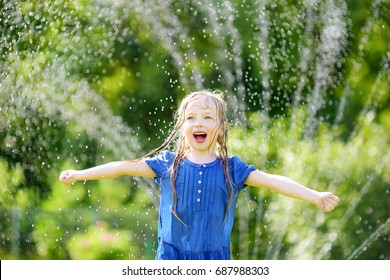 Adorable Little Girl Playing With A Sprinkler In A Backyard On Sunny Summer Day. Cute Child Having Fun With Water Outdoors. Funny Summer Games For Kids.