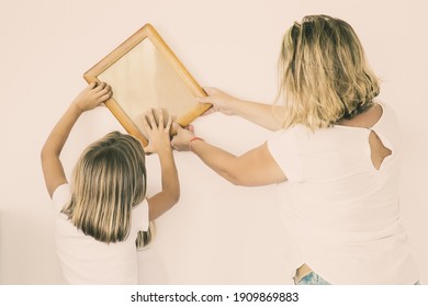 Adorable Little Girl Putting Photo Frame On White Wall With Help Of Mom. Cute Daughter And Blonde Mother Hanging Blank Picture. Family Decorating Room Together. Relocation And Moving Day Concept