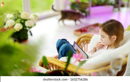 Adorable Little Girl Reading An Ebook In White Living Room On Beautiful Summer Day. Smart Schoolgirl Doing Her Homework At Home After School.