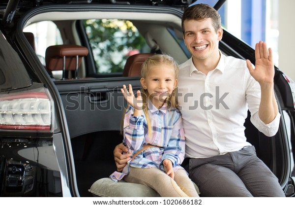 Adorable little girl sitting in an open trunk\
of a car with her cheerful father waving to the camera happily\
automobile cars travelling journey trip vacation family bonding\
children kids\
parenthood