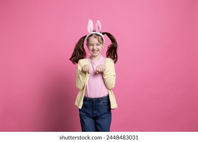 Adorable niña que pasa las vacaciones de Pascua saltando como un conejo para la cámara mientras lleva orejas de conejo. Encantado joven feliz jugando y riéndose en el fondo rosa.