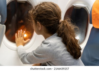 Adorable Little Girl Traveling By Plane. Kid Sits At The Window Of The Airplane And Looks Out The Window From The Outside. Night Flight With Children