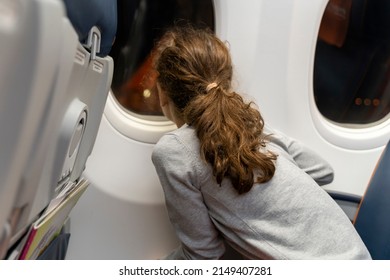 Adorable Little Girl Traveling By Plane. Kid Sits At The Window Of The Airplane And Looks Out The Window From The Outside. Night Flight With Children