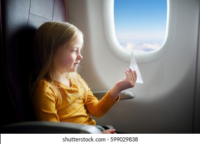 Adorable Little Girl Traveling By An Airplane. Child Sitting By Aircraft Window And Playing With Paper Plane.