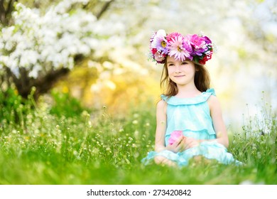 Adorable Little Girl Wearing Flower Crown Sitting In Blooming Cherry Garden On Beautiful Spring Day