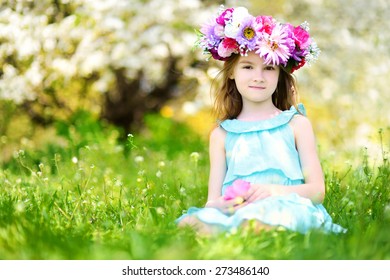 Adorable Little Girl Wearing Flower Crown Sitting In Blooming Cherry Garden On Beautiful Spring Day