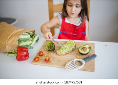 Adorable Little Girl At White Table Spreading Avocado On Slice Of Bread. Cute And Happy Young Child Cutting Vegetables On Cutting Board. Kids Preparing Food