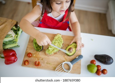 Adorable Little Girl At White Table Spreading Avocado On Slice Of Bread. Cute And Happy Young Child Cutting Vegetables On Cutting Board. Kids Preparing Food