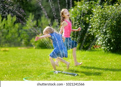 Adorable Little Girls Playing With A Sprinkler In A Backyard On Sunny Summer Day. Cute Children Having Fun With Water Outdoors. Funny Summer Games For Kids.