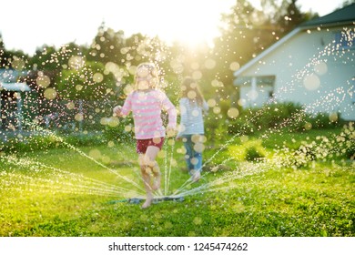 Adorable Little Girls Playing With A Sprinkler In A Backyard On Sunny Summer Day. Cute Children Having Fun With Water Outdoors. Funny Summer Games For Kids.