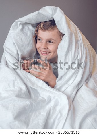 Adorable little kid with blond hair wrapped in white duvet holding glass of milk and looking away at home in morning