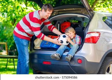 Adorable Little Kid Boy Sitting In Car Trunk Just Before Leaving For Summer Vacation. Dad Packing Suitcases. Happy Family Going On Long Journey.