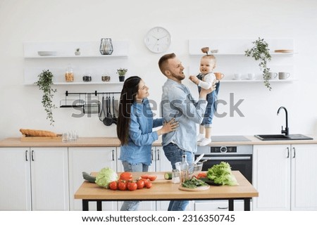 Adorable little lady in denim clothes laughing with caring parents while dad holding baby daughter in arms. Happy spouses and kid building lifelong memories while upholding family culinary tradition.
