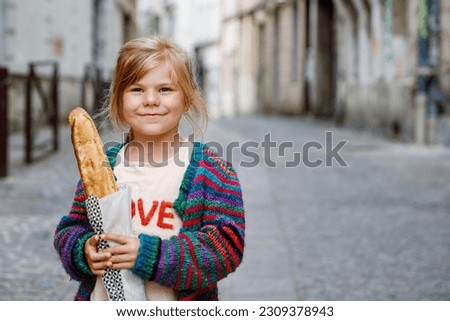 Adorable little preschool girl with fresh French baguette on the street side of the city. Happy small child in Paris, France.