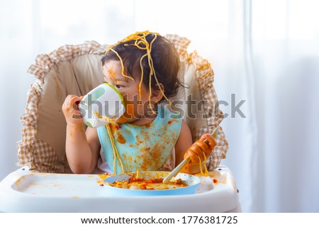 Adorable little toddler girl or infant baby drink water after eat delicious spaghetti food on chair. Funny cute infant girl get thirsty. Lovely mix race daughter get dirty. Kid hold spoon upside down