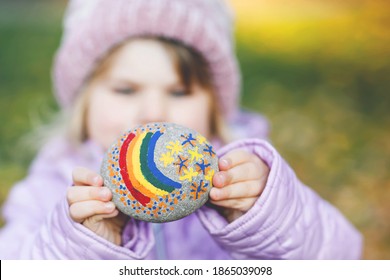 Adorable Little Toddler Girl With Painted Rainbow On Stone During Pandemic Coronavirus Quarantine, Outdoors. Child Painting Rainbow