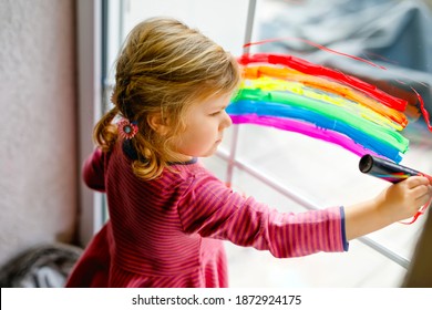 Adorable Little Toddler Girl With Rainbow And Heart Painted With Colorful Window Color During Pandemic Coronavirus Quarantine. Child Painting Rainbows Around The World With The Words Let's All Be Well