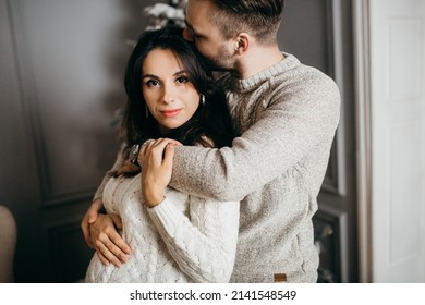 Adorable marriage couple stand near Christmas tree at home, caring husband hug pregnant wife, young family excited about parenthood, enjoy happiness, winter holidays concept. High quality photo - Shutterstock ID 2141548549
