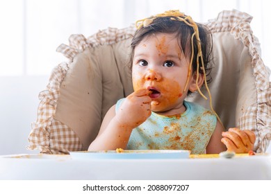 Adorable Messy Little Child Girl Hungry Use Hand Eating Spaghetti Sitting In High-powered Chair At Home. Toddler Child With Tomato Sauce Making Mess Face Looking At Parent. Self-feeding Concept
