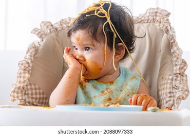 Adorable Messy Little Child Girl Hungry Use Hand Eating Spaghetti Sitting In High-powered Chair At Home. Toddler Child With Tomato Sauce Making Mess Face Looking At Parent. Self-feeding Concept