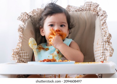 Adorable Messy Little Child Girl Hungry Use Hand Eating Spaghetti Sitting In High-powered Chair At Home. Toddler Child With Tomato Sauce Making Mess Face Looking At Parent. Self-feeding Concept