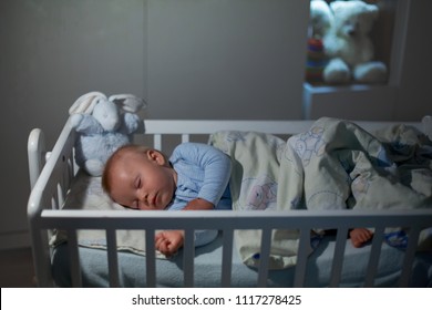 Adorable Newborn Baby Boy, Sleeping In Crib At Night. Little Boy In White Pajamas Taking A Nap In Dark Room. Bedroom Interior With Lamp And Teddy Bear In The Background