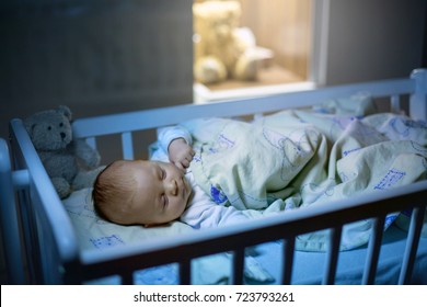 Adorable Newborn Baby Boy, Sleeping In Crib At Night. Little Boy In White Pajamas Taking A Nap In Dark Room. Bedroom Interi?r With Lamp And Teddy Bear In The Background 