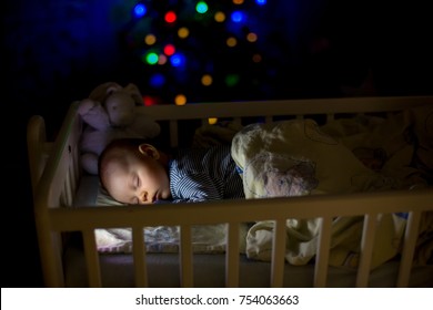 Adorable Newborn Baby Boy, Sleeping In Crib At Night. Little Boy In Blue Striped Pajamas Taking A Nap In Dark Room, Christmas Decoration In The Room, Winter Time, Snowing Outdoors