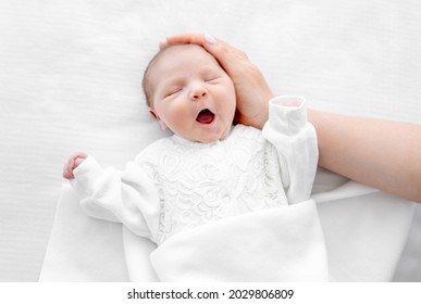Adorable newborn baby girl wearing white costume lying in the bed at home with daylight, yawning during sleeping and her mother hand cares about her. Portrait of cute infant child and parent love