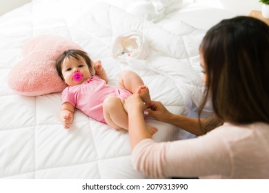 Adorable Newborn Baby Using A Pacifier And Wearing A Pink Onesie. Loving Mom Dressing Her Baby In Bed In The Morning