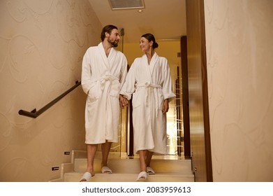 Adorable Newlywed Couple In Love, Dressed In White Bathrobes, Enjoying Relaxation Together In Luxury Spa During Their Honeymoon, Holding Hands As They Walk Down The Stairs Of The Wellness Spa Complex