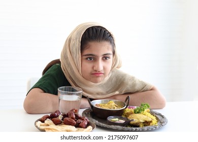 Adorable Pakistani Muslim Girl With Beautiful Eyes Sitting At Kitchen Table, Kid Wearing Hijab Bored (Anorexia) With Traditional Islamic Halal Food On White Background.