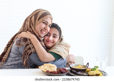 Adorable Pakistani Muslim Girl Sits At Kitchen Table With Traditional Islamic Halal Food, Kid With Hijab Hugs Mother, Warm Love In Family Of Mom And Daughter, Happy Islamic Family On White Background.