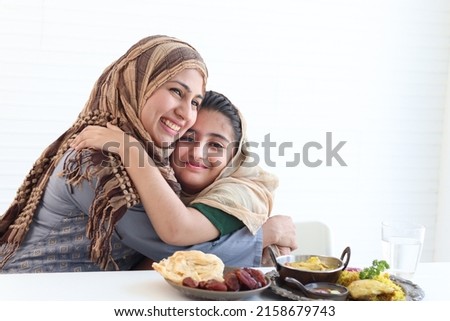 Adorable Pakistani Muslim girl sits at kitchen table with traditional Islamic halal food, kid with hijab hugs mother, warm love in family of mom and daughter, happy Islamic family on white background.