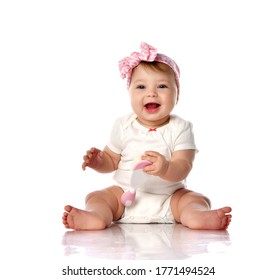 Adorable Positive Little Baby Girl In White Cotton Body, Hair Bow Decoration And Barefoot Sitting On Floor, Smiling And Holding Toy In Hand Over White Background. Happy Childhood Concept