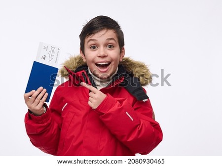 Adorable preadolescent boy, cheerful school child in bright red warm parka points with his finger at a passport with a ticket and boarding pass, isolated over white background with copy ad space