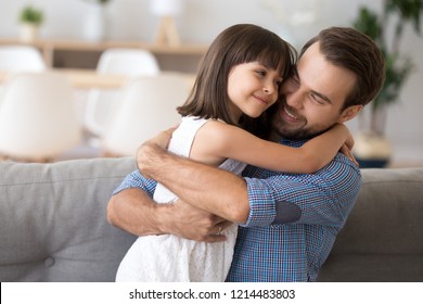 Adorable preschool daughter wearing white dress hugging loving father looking at him with love and tenderness. Multi-ethnic diverse friendly family on couch at modern home spending free time together