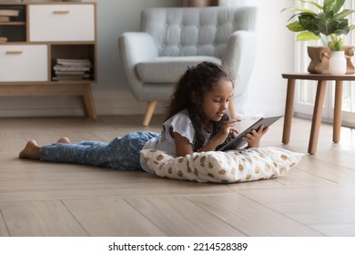 Adorable preschooler little Indian girl lying on warm floor using digital tablet in cozy living room, play videogames, watch favourite vlog, spend time at home. Gen Z use modern wireless technology - Powered by Shutterstock