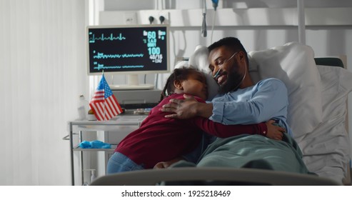 Adorable Preteen Black Girl Visiting Sick Dad In Hospital. Smiling African Patient Resting In Bed At Hospital Ward Meeting And Hugging Cute Daughter