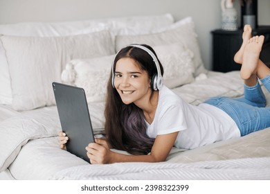 Adorable pre-teen girl in wireless headphones lying on bed with digital tablet device. Young generation and modern tech overuse, gadget usage for fun, hobby and leisure on weekend at home - Powered by Shutterstock