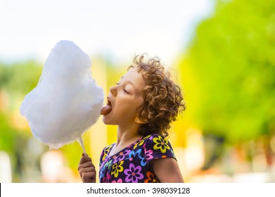 Adorable And Pretty Curly Little Girl Eating White Sweet Cotton Candy. Happy Child, Kid Eating Candy-floss With Emotions In The Park At Summer Or Spring.