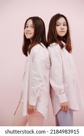 Adorable Pretty Twin Girls Standing Back To Back, Both Looking Over The Shoulder. Over Pink Background.